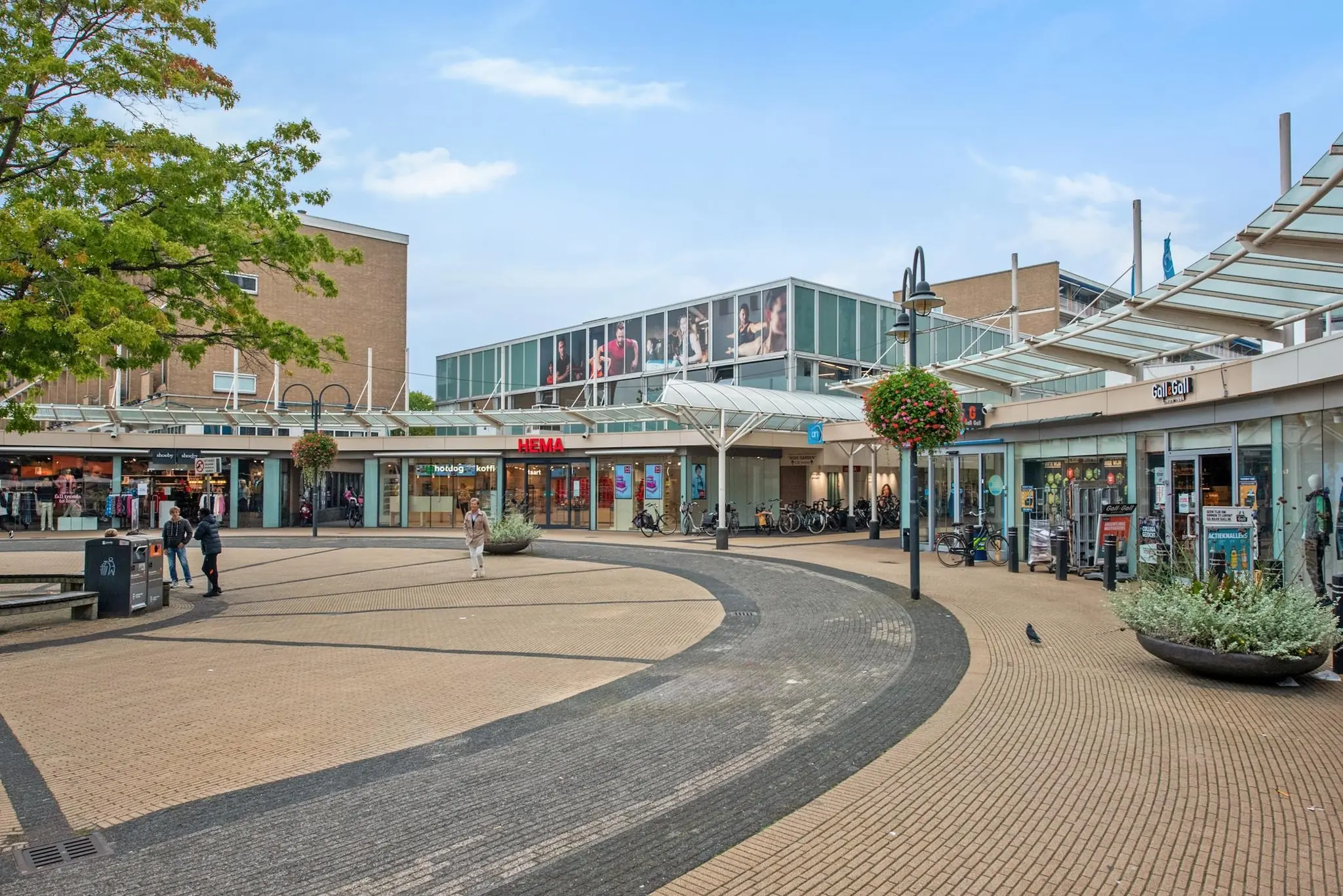 Winkelcentrum aan de Koningin Julianalaan met diverse winkels zoals HEMA en Call & Golf, en een open plein met voetgangers en fietsen.